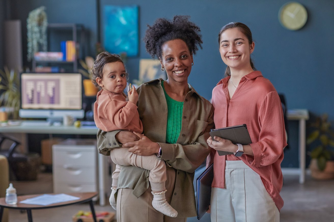 Two smiling young adults and a baby pose in a modern office environment. One adult holds the baby, who is looking towards the viewer, while the other adult holds a tablet and folder.