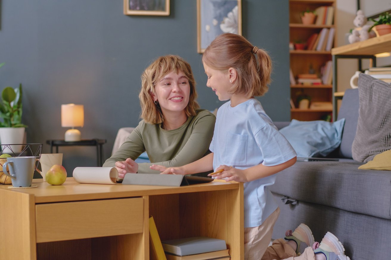 A smiling young adult sits at a wooden table, looking warmly at a child who stands nearby with a pencil, as they both engage with a tablet and papers in a comfortable room.