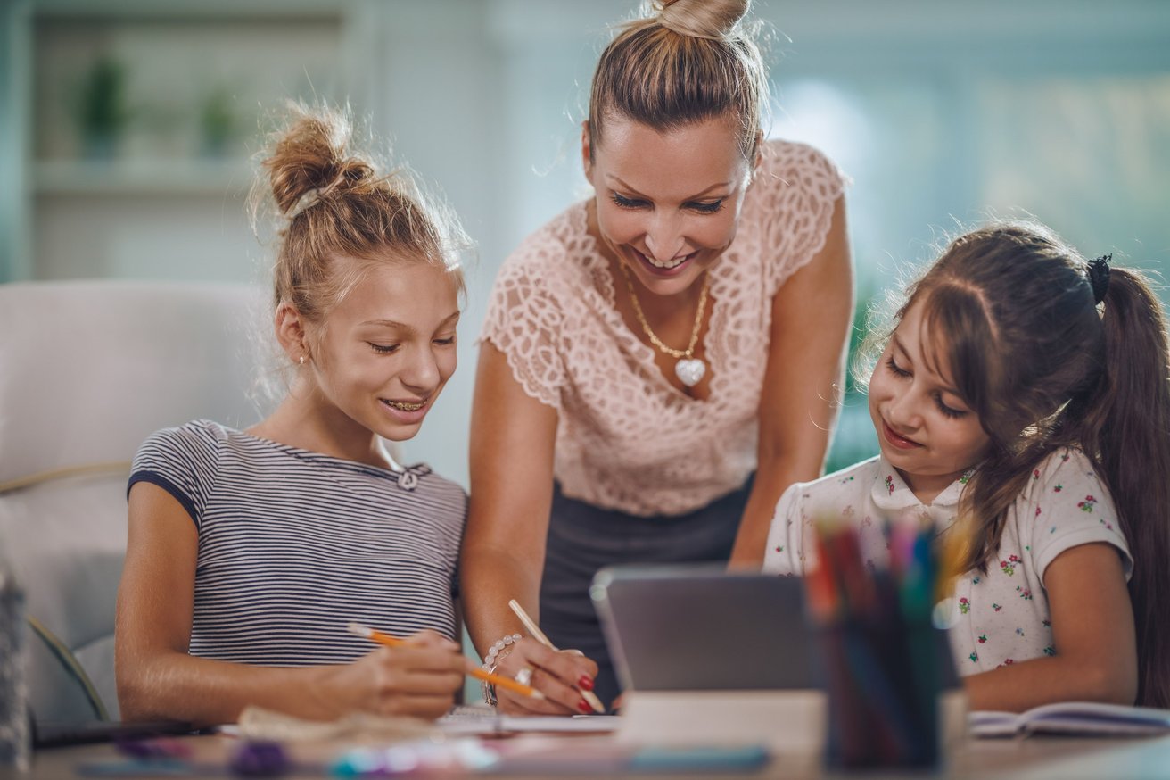 An adult woman smiles as she guides a pre-teen and a younger child in a learning activity at a table. They are writing and looking at a tablet in a bright home setting.
