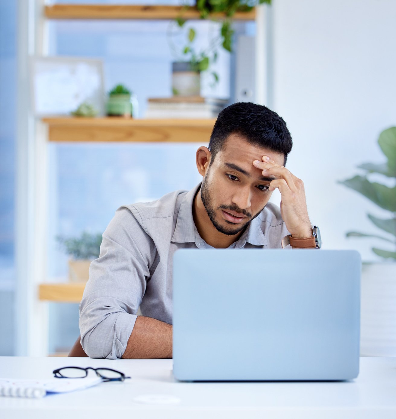 An adult male sits at a desk, looking intently at a laptop screen with one hand on his forehead, conveying a stressed or focused mood in an office setting.