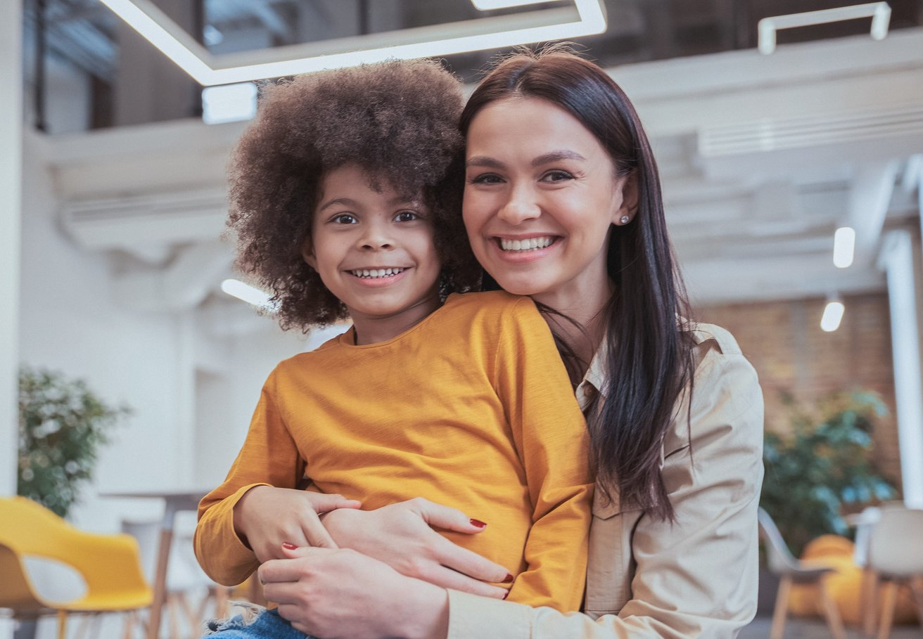 An adult woman warmly embraces a smiling young child, both looking directly at the camera. They are in a bright, modern indoor setting, conveying a mood of happiness and connection.