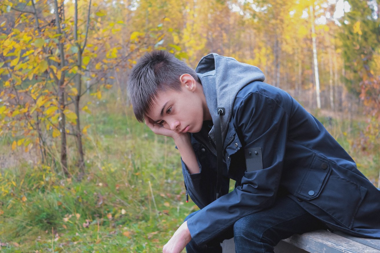 A young male teenager sits alone on a wooden bench outdoors amidst autumn foliage, looking down with a somber and pensive expression, his head resting on his hand.