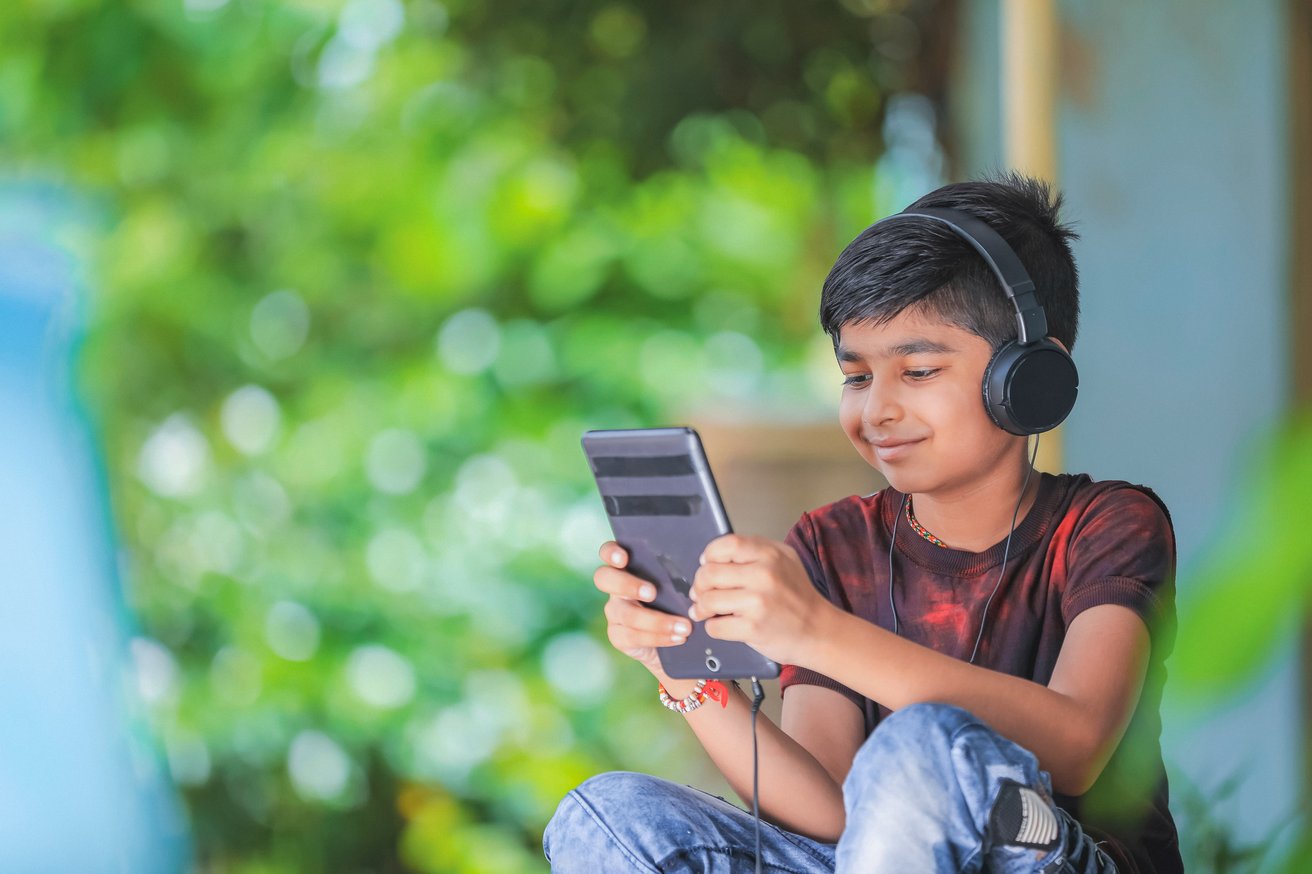 A young boy wearing black headphones is smiling while looking at a digital tablet or large smartphone. He is seated outdoors, with a blurred green leafy background, appearing calm and engaged with his device.
