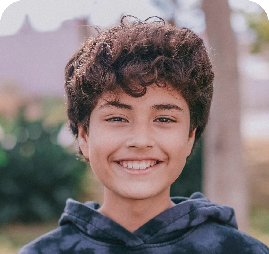 A happy child with curly brown hair and bright brown eyes smiles directly at the camera, wearing a dark hoodie. The background is a softly blurred outdoor setting.