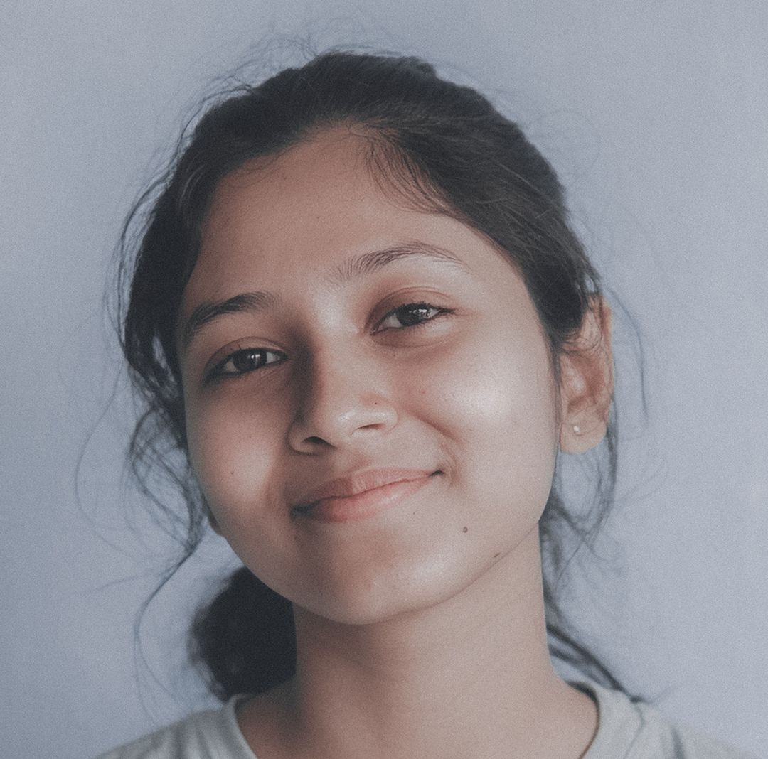 A close-up portrait of a smiling young woman with dark hair pulled back. She looks directly at the camera with a gentle, approachable expression against a plain, light-colored background.