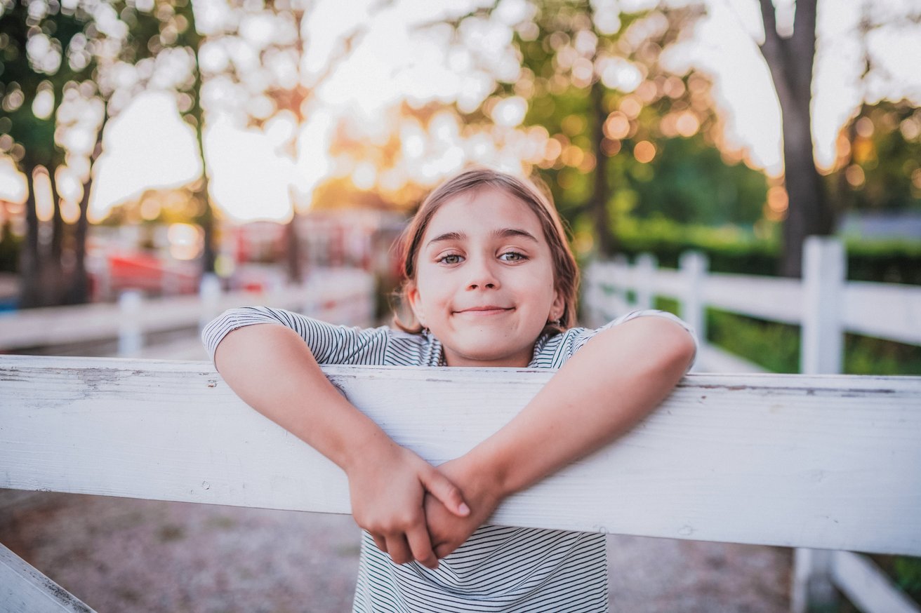 A happy young child leans on a white wooden fence outdoors, smiling gently at the viewer. The background is softly blurred with green trees and warm, bright sunlight.