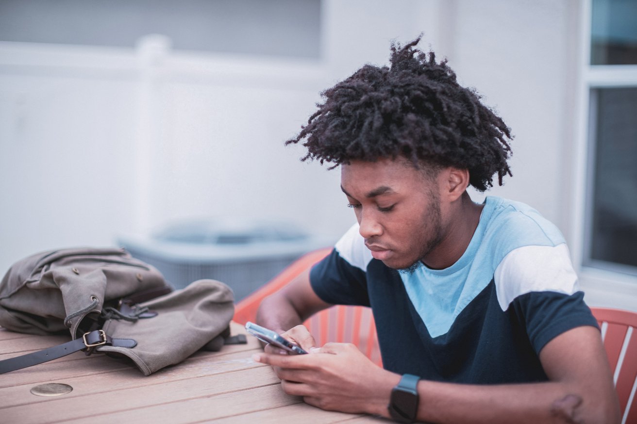 A young man, appearing to be a teenager, sits at a wooden table outdoors, deeply focused on his smartphone. A canvas backpack rests on the table next to him.