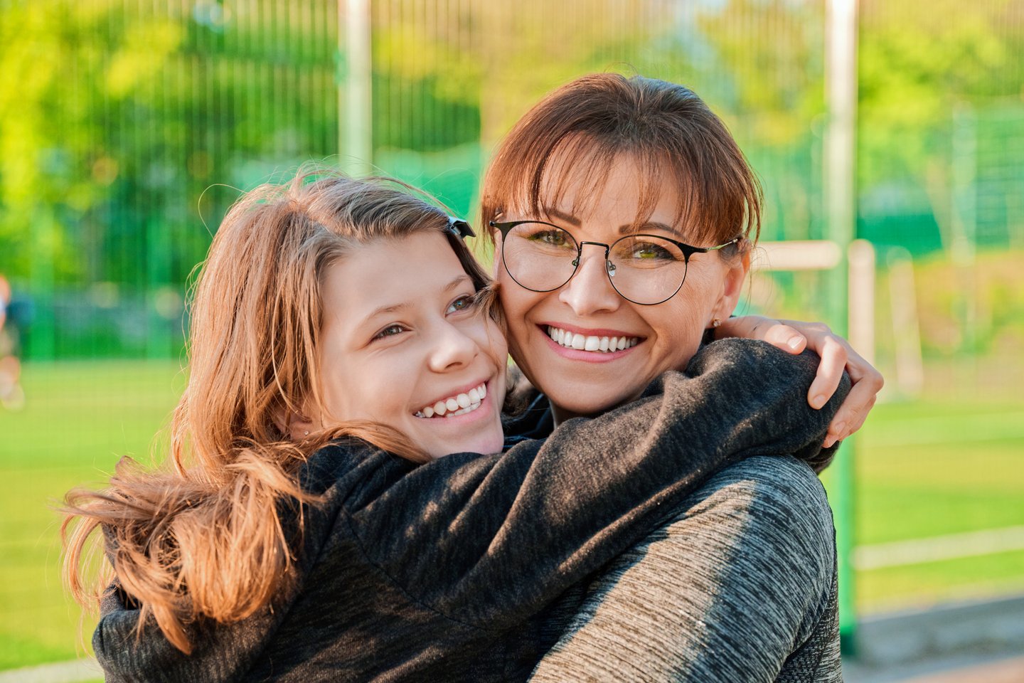 An adult woman wearing glasses is embracing a teen girl outdoors, with both smiling warmly. The background is a blurred green field.