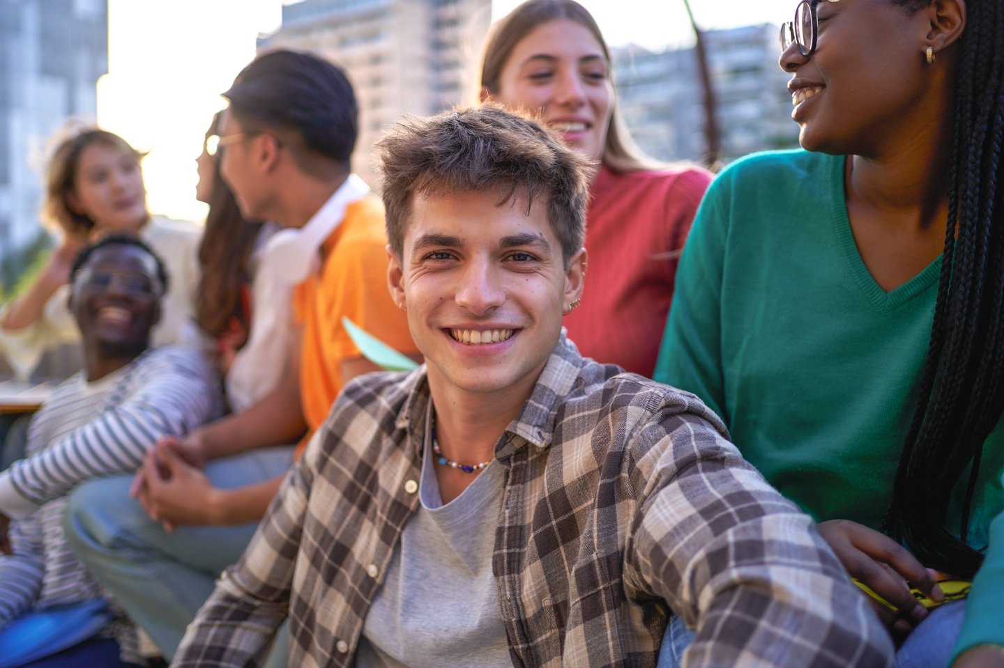 A smiling young man in a plaid shirt looks at the viewer, surrounded by a group of happy teens and young adults in an outdoor urban setting.