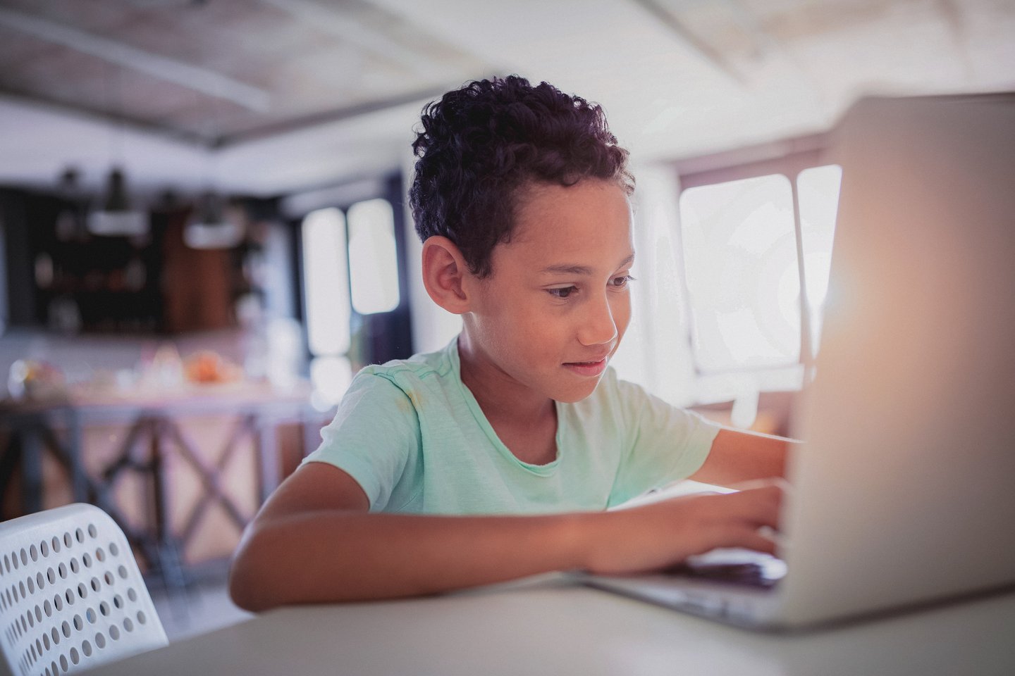 A young boy with curly hair is focused on typing on a laptop, which is placed on a light-colored table in an indoor setting. He appears engaged with the screen.