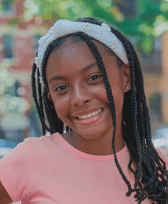 A smiling pre-teen girl with long braided hair and a white daisy-decorated headband looks directly at the viewer outdoors. She appears happy and friendly.
