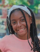 A smiling young girl with long braided hair and a white daisy headband poses for a close-up portrait outdoors. She looks happy and friendly.