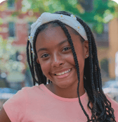 A happy young girl with long braids and a white daisy headband smiles brightly at the camera in an outdoor setting with blurred greenery and buildings.