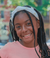 A young girl with long braided hair and a white headband adorned with daisies smiles brightly while looking directly at the camera. She is outdoors, and her expression conveys happiness and approachability.