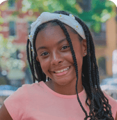 A smiling pre-teen girl with long braided hair and a white daisy headband stands outdoors in a bright, positive mood.