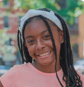A smiling pre-teen girl with long braided hair and a white daisy headband looks directly at the camera. She appears confident and happy in an outdoor setting with a blurred background.