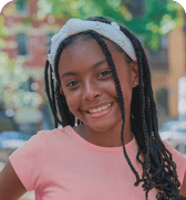 A cheerful young girl with braided hair and a daisy-decorated headband smiles broadly, captured in a vibrant outdoor portrait.