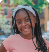 A young girl with long dark braided hair and a white daisy-decorated headband smiles brightly at the viewer. She is wearing a pink t-shirt and is captured in an outdoor setting with a blurred background.