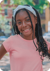 A young girl with long braided hair and a white daisy-decorated headband smiles brightly at the camera. She is outdoors, wearing a pink t-shirt, with a blurred natural background.
