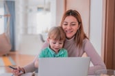 An adult woman and a young child are smiling and looking at a laptop together at a table. The woman holds a pen and a notebook, suggesting an interactive or learning moment in a bright, indoor setting.