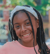 A joyful young girl with braided hair and a white daisy headband smiles brightly at the viewer, set outdoors with a blurred background.