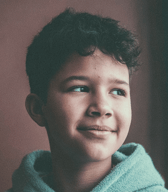 A young boy with curly hair, wearing a blue hooded top, looks calmly to the side with a gentle smile. He is captured in a close-up portrait indoors.
