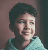 A young boy, around 8-12 years old, is shown in a close-up portrait, looking thoughtfully to the side with a gentle, hopeful smile, wearing a soft, light teal top.