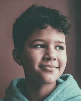 A smiling Hispanic child with curly hair looks thoughtfully to the side while wearing a comfortable hooded garment. The close-up portrait conveys a calm and hopeful mood.