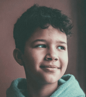 A young boy with dark curly hair looks gently to the side with a soft, pensive smile. He is wearing a light blue hooded garment, captured in a close-up portrait against a muted background.