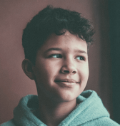 A young boy with dark curly hair looks off to the right with a soft, gentle smile against a blurred reddish-brown background. He is wearing a light blue textured hoodie.