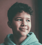 A young boy with dark, curly hair looks thoughtfully to his right with a gentle smile, wearing a light blue hoodie indoors.