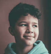 A young boy with curly dark hair smiles softly and looks contemplatively to the side. He is wearing a soft blue hoodie in this close-up portrait, conveying a sense of calm.