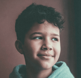 A young boy with dark, curly hair smiles gently while looking thoughtfully to the side. He is positioned indoors against a plain background, exuding a calm and content mood.