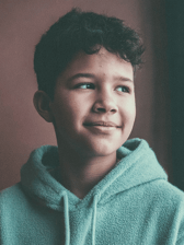 A pre-teen boy with curly dark hair, wearing a teal hoodie, looks to the side with a gentle smile. He appears calm and content in an indoor setting.