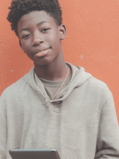 A male teenager with short, curly hair smiles faintly at the camera while holding a tablet. He stands casually against a vibrant orange wall.