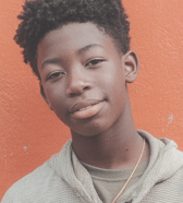 A young teen boy with dark, curly hair looks directly at the camera with a gentle smile, posing against a vibrant orange wall. He wears a light-colored hooded top, conveying an approachable and calm mood.