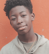 A teen boy with short, textured hair smiles gently while looking directly at the camera. He wears a casual hoodie and stands against a bright orange wall, conveying a calm and friendly mood.