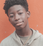 A smiling teen boy with short curly hair is captured in a close-up portrait, looking calmly at the viewer against a plain orange background.