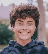 A smiling pre-teen boy with curly brown hair looks directly at the viewer, radiating a positive and friendly mood. He is outdoors, wearing a dark hoodie.