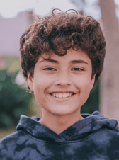 A young boy with curly brown hair smiles brightly directly at the camera. He appears happy and friendly, likely in an outdoor setting with a blurred natural background.