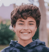 A pre-teen boy with curly brown hair smiles brightly at the camera, wearing a dark hoodie. The background is blurred, suggesting an outdoor setting and a positive, confident mood.