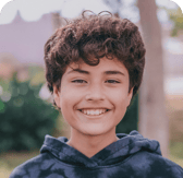 A young boy with curly brown hair and expressive dark eyes smiles broadly at the viewer. He is wearing a dark, patterned hoodie, and the background is a softly blurred outdoor setting with natural light, conveying a sense of happiness and confidence.