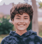 A smiling child with short, curly brown hair, wearing a dark tie-dye hoodie, looks directly at the camera. The background is a softly blurred outdoor setting with trees and greenery, suggesting a bright and positive mood.