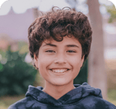 A happy child with curly brown hair and bright brown eyes smiles directly at the camera, wearing a dark hoodie. The background is a softly blurred outdoor setting.