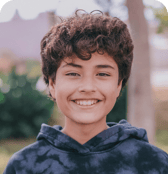 A young boy with dark curly hair smiles confidently at the camera. He is wearing a tie-dye hoodie and is outdoors with a softly blurred natural background.
