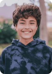 A young boy with curly brown hair is smiling brightly at the camera. He is wearing a dark blue tie-dye hoodie and is outdoors with a blurred natural background.