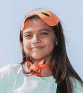 A smiling young girl with long brown hair looks directly at the camera, wearing an orange superhero mask on her forehead and a matching ribbon around her neck. She appears happy and playful outdoors under a clear blue sky.