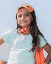 A confident young girl wears an orange superhero mask and cape, smiling broadly with her hands on her hips. She stands outdoors against a bright, light background, embodying a sense of strength and playfulness.