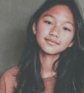 A young teen girl of East Asian ethnicity with long dark hair smiles gently, looking directly at the camera in a close-up portrait. She conveys a calm and friendly mood against a simple background.