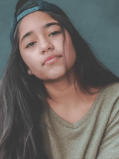 A young person, likely a teenager, with long dark hair and a backward dark cap, looks directly at the camera with a contemplative expression. They are wearing a casual olive-green v-neck top against a plain background.